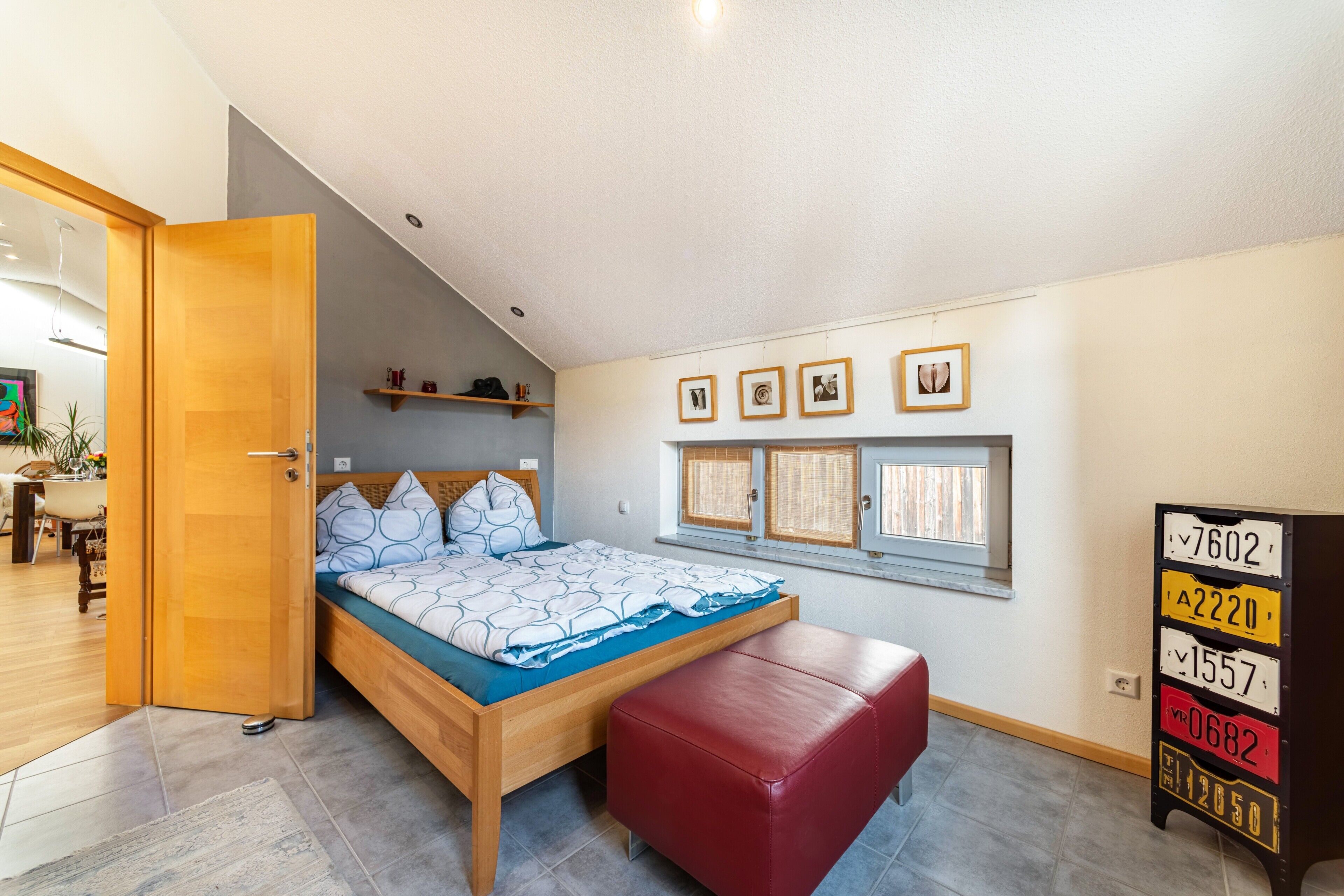 Ein Schlafzimmer mit einem Bett, einer roten Bank, einer Holztür, einem Regal mit Gegenständen sowie einer Wand mit gerahmten Bildern und Nummernschildern.