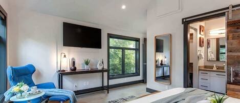 A modern bedroom with a bed, a desk, a chair, a TV, and a bathroom visible through an open door.