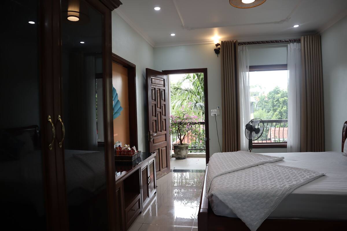 A bedroom with a large bed, a wooden wardrobe, a desk, and a balcony with a view of greenery.