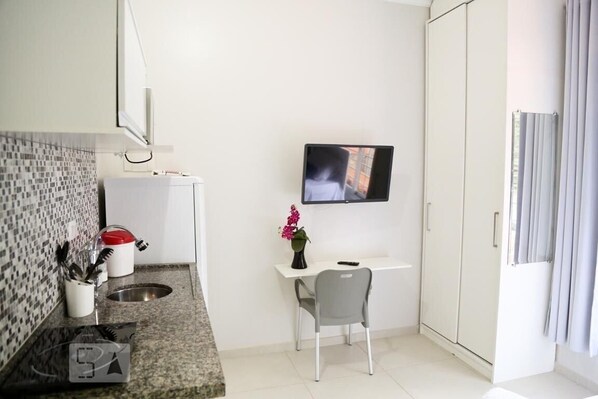 A small, modern kitchen with a microwave, sink, and a small table with a chair. There is a TV mounted on the wall and a window with curtains. - Vila Nina (São Paulo)