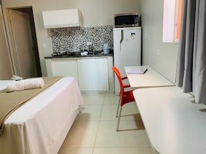 Una habitación de hotel compacta con kitchenette, una cama con cubrecama blanco, una silla roja y una mesa con un cuaderno.