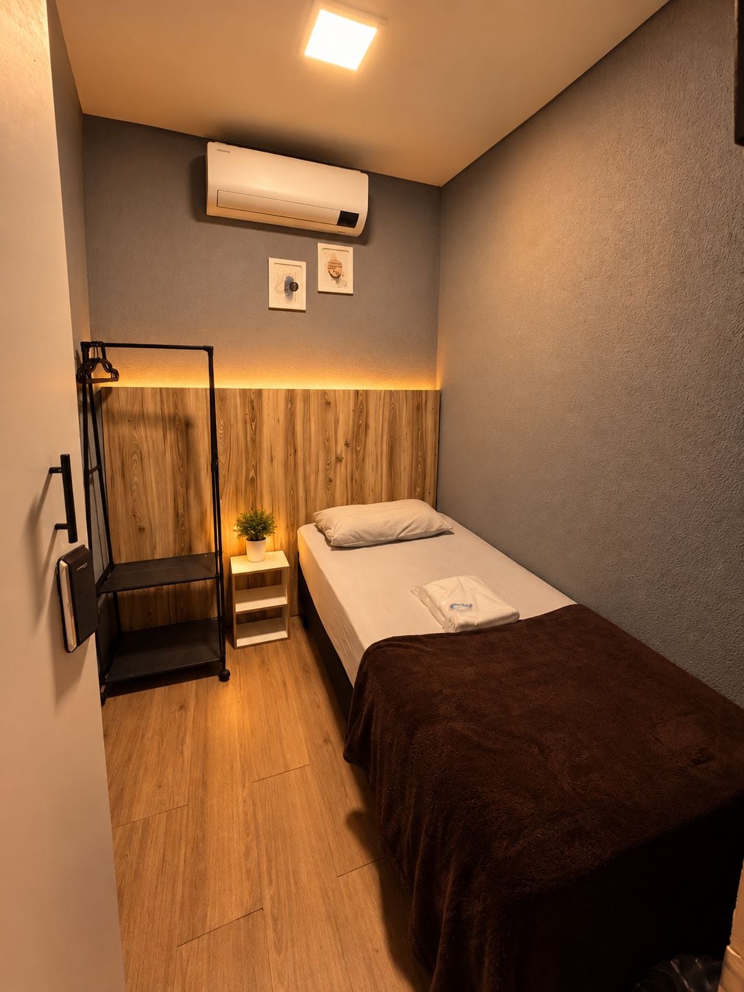 A compact hotel room with a bed, bedside table, a small plant, a wall-mounted air conditioner, and a wooden accent wall.