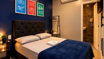 Un dormitorio con una cama grande, una mesita de noche con lámpara, aire acondicionado de pared y un cartel enmarcados con palabras relacionadas con viajes.