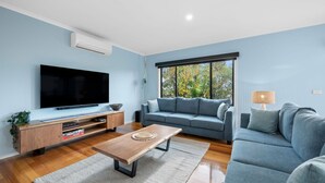 Ein modernes Wohnzimmer mit Flachbildfernseher, Couchtisch aus Holz, grauem Sofa und großem Fenster mit Blick auf Bäume.