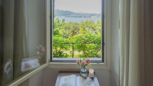Una ventana con vistas a un lago y vegetación, un florero con flores, y un libro abierto sobre una pequeña mesa.