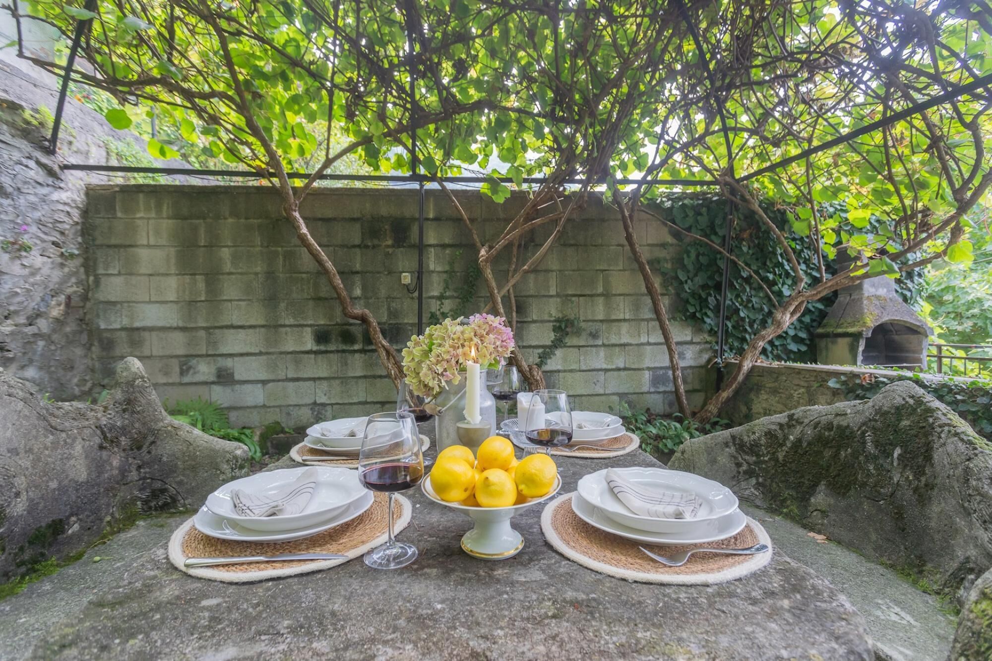 Une table en pierre dressée pour un repas, avec des assiettes, des couverts, des verres et un saladier rempli de citrons.