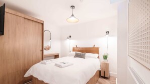 Quarto bem arrumado com guarda-roupa de madeira, cama com roupa de cama branca, duas luminárias de cabeceira e uma lâmpada no teto.