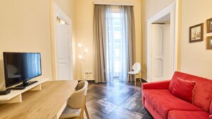 Ein Wohnzimmer mit Fernseher, einem roten Sofa, einem Esstisch mit Stühlen und einem Fenster mit Vorhängen.