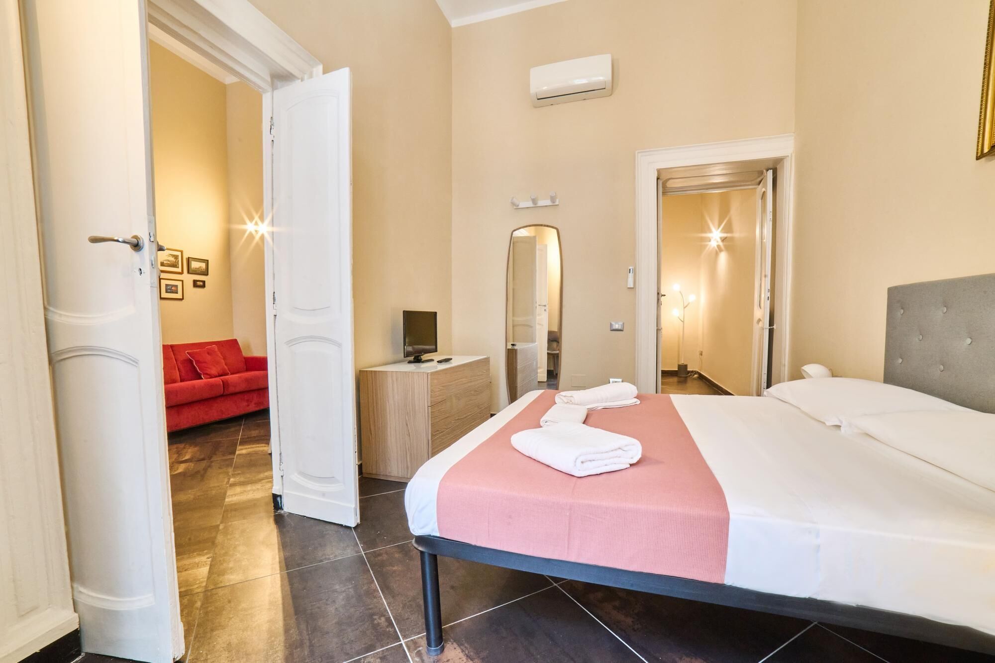 Quarto de hotel com uma cama, uma mesinha de cabeceira, um espelho e um sofá vermelho no quarto adjacente.