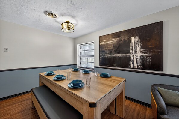 A dining area with a wooden table set for four, a bench, a large abstract painting on the wall, and a window with blinds.