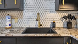 Eine Küche mit einem schwarzen Spülbecken, einem Wasserhahn und einer Rückwand aus sechseckigen Fliesen. Es sind Schränke mit goldenen Griffen und eine Seifenspenderdose zu sehen.