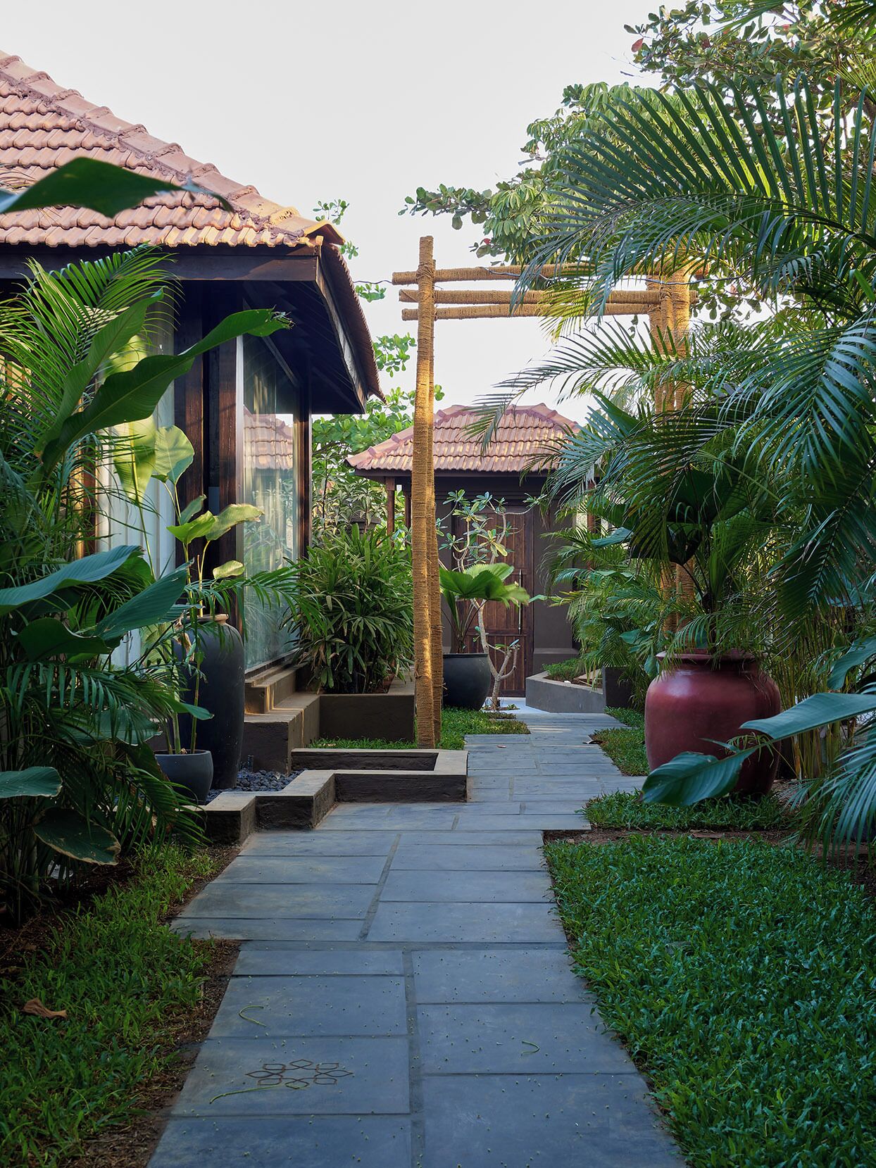 A pathway leading to a building with a tiled roof, surrounded by lush greenery and large potted plants.