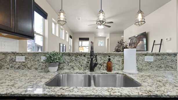A kitchen with granite countertops, a stainless steel sink, pendant lights, and a ceiling fan.