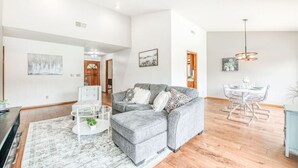 Een ruime woonkamer met een grijze hoekbank, een glazen salontafel en een eethoek met een witte tafel en stoelen.
