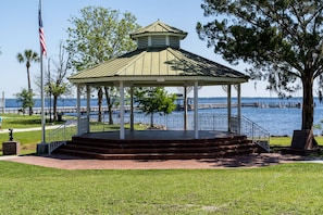 Un pabellón con techo metálico y columnas blancas, ubicado en una zona verde con vistas a un cuerpo de agua y un muelle al fondo.