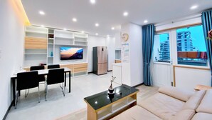 Ett modernt vardagsrum med en platt-TV, en soffa, en matplats med stolar, ett köksområde och ett fönster med utsikt över staden.