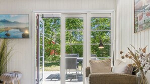 A room with a wicker sofa, a dining table, and a large sliding glass door leading to an outdoor area with a view of trees and a building. - "Blakke" - 250m from the sea by Interhome (Nexø)