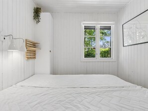 A bedroom with a bed, a window with a view of greenery, a wall-mounted light, and a framed picture.