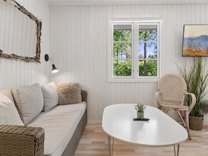 A room with a white wicker sofa, a round white table, wicker chairs, a window with a view of trees, and a wall-mounted picture.