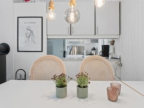 A modern kitchen with white cabinets, a framed artwork, wicker chairs, and a dining table with potted plants and glassware.