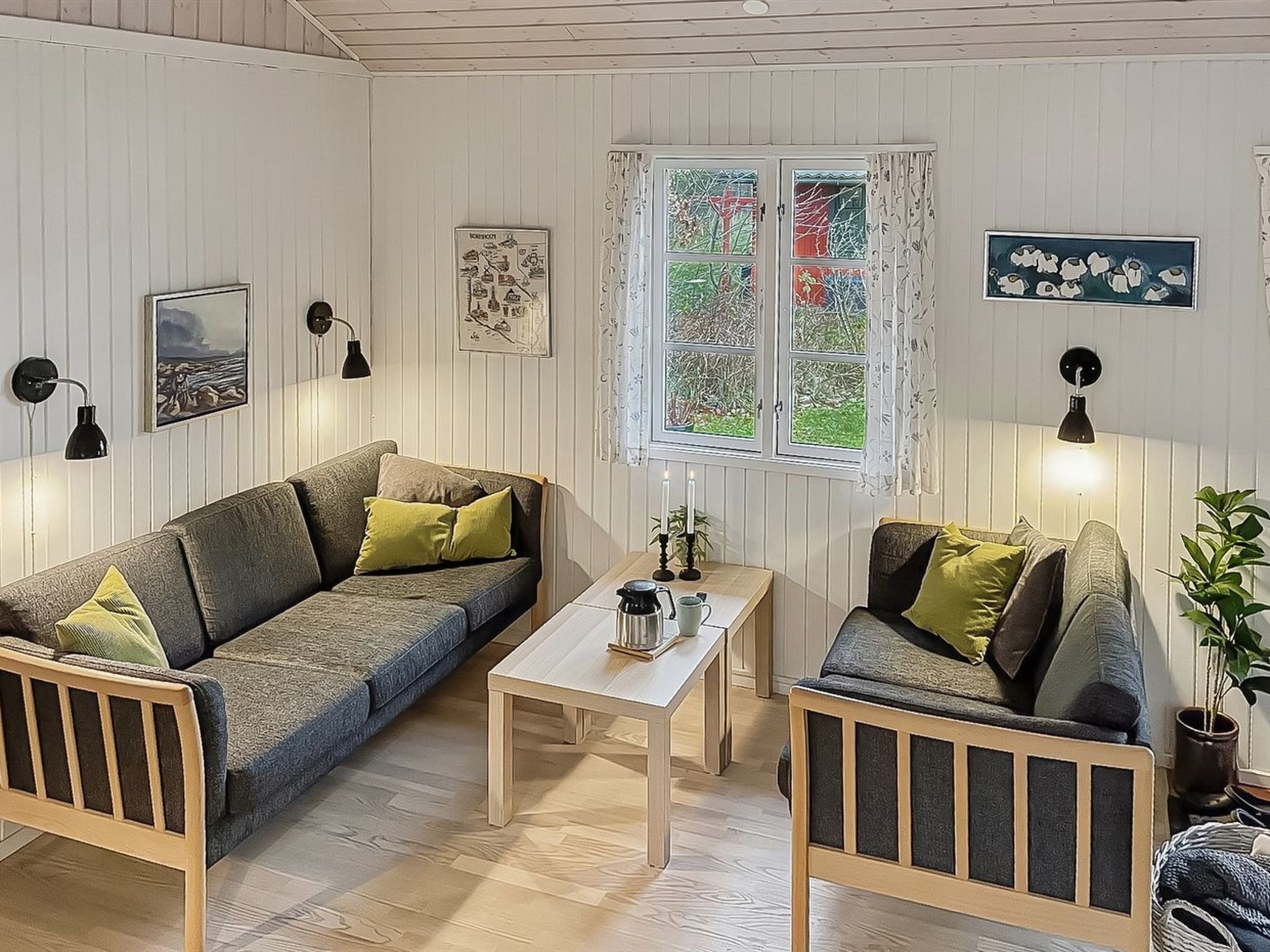 Ein gemütliches Wohnzimmer mit einer Couch, einem kleinen Tisch und einem Sessel. Ein Fenster mit Vorhängen, eine Lampe und Wandbilder sind zu sehen.