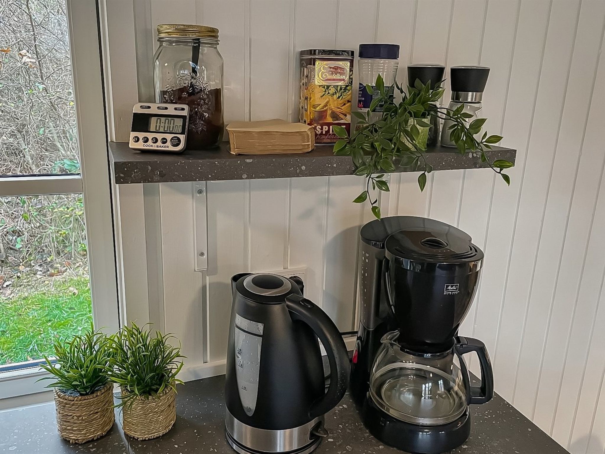 A kitchen with a coffee maker, a blender, a jar of jam, and a shelf with various items.
