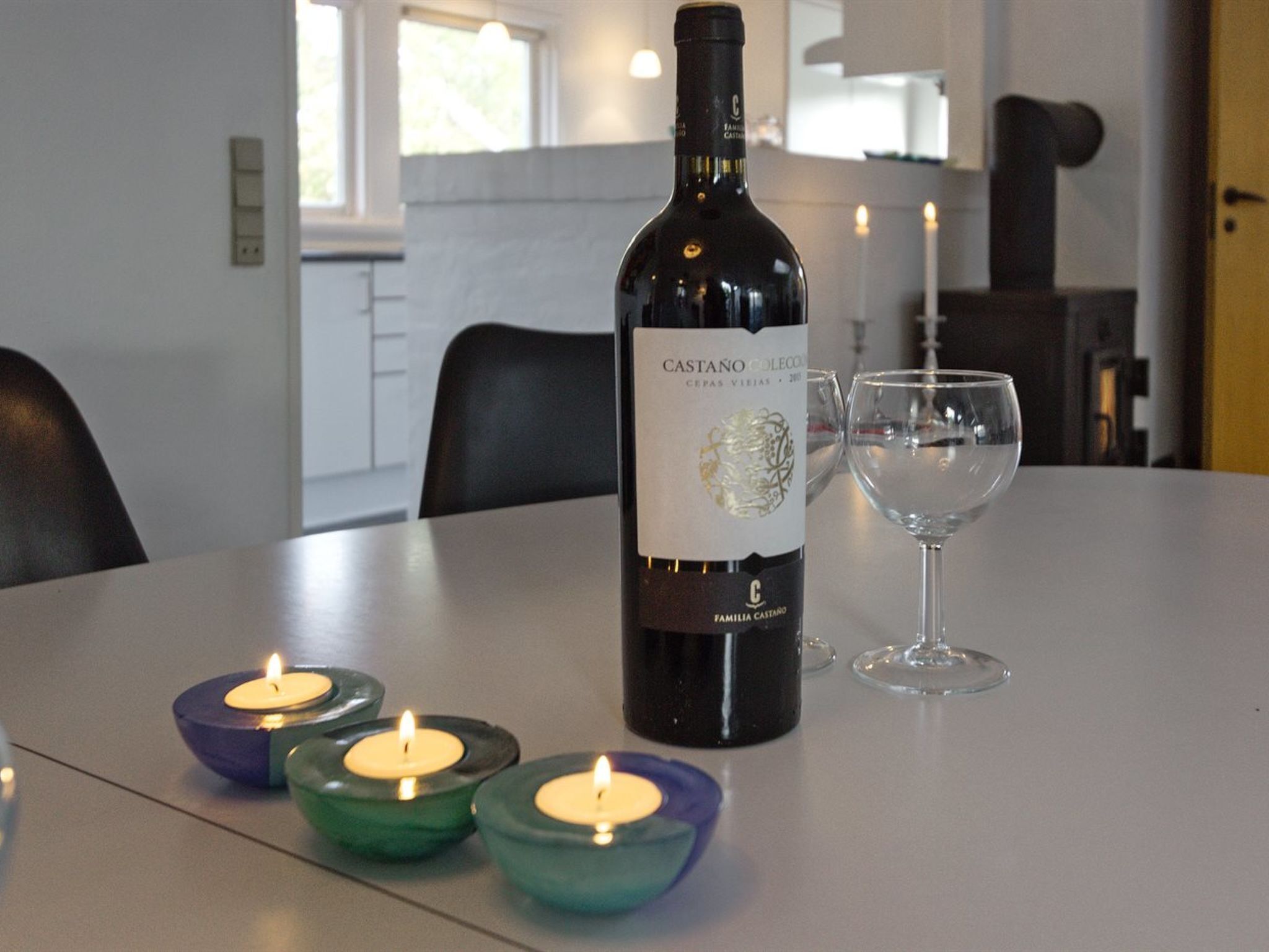 A bottle of Castano Coleccion wine, two glasses, and three lit candles on a table in a modern kitchen.