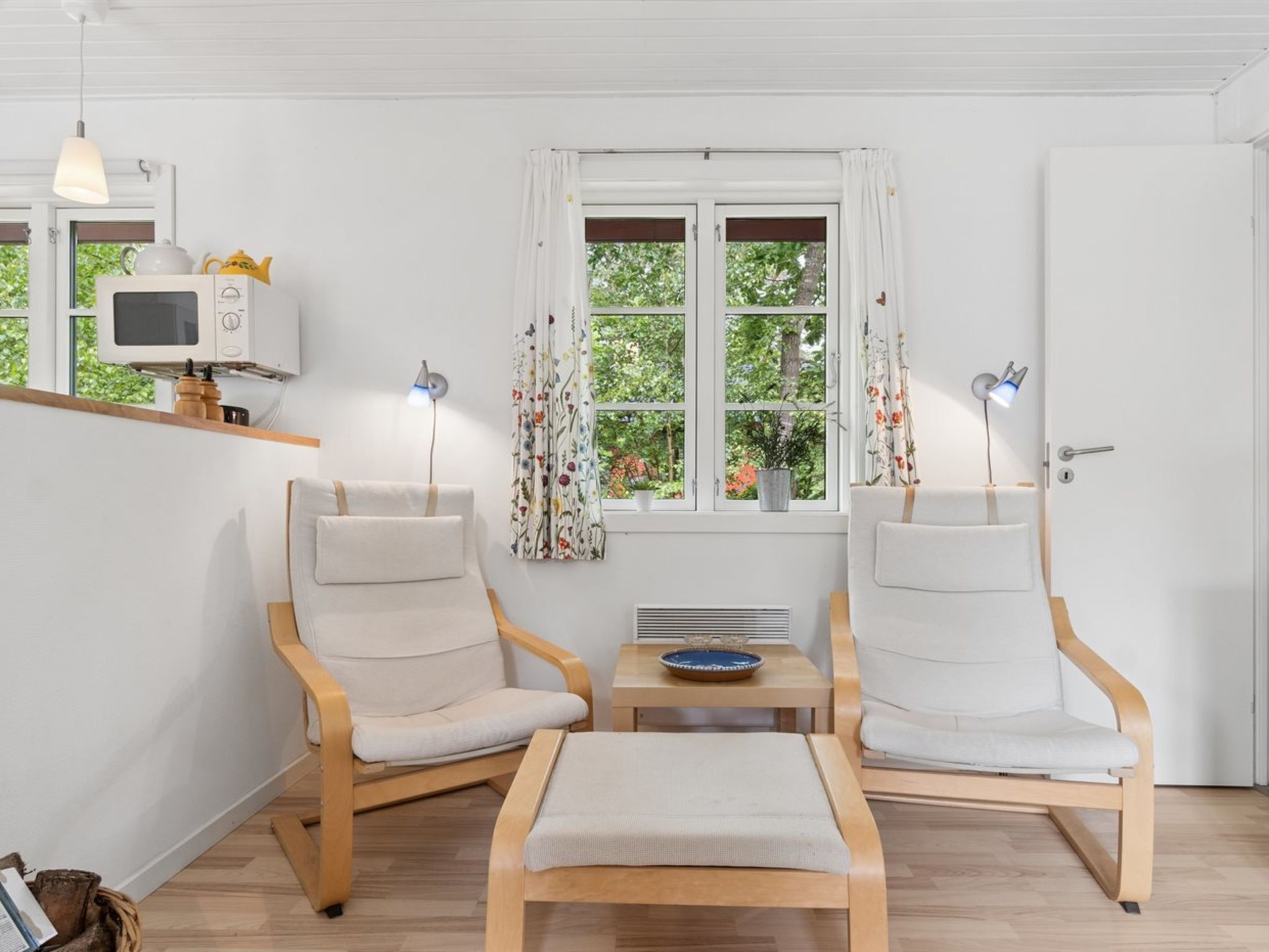 Una habitación con dos sillas de madera, una mesita, microondas, lámpara y ventana con cortinas.