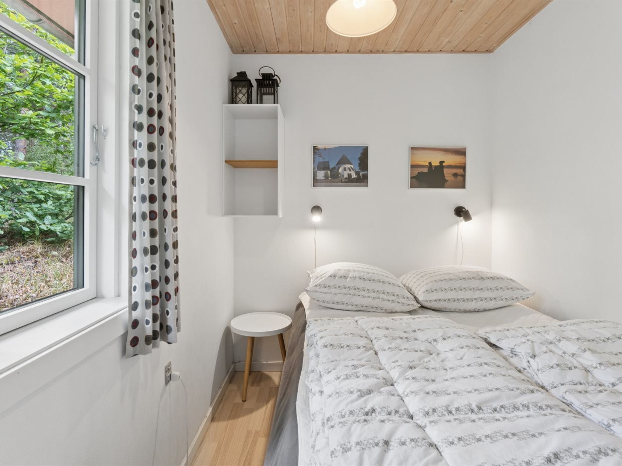 A bedroom with a bed, a small bedside table, a window with curtains, and two wall-mounted lights.