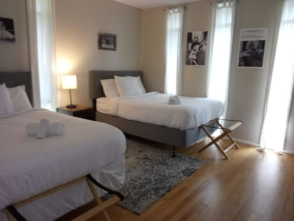 Een hotelkamer met twee bedden, een nachtkastje met een lamp, een klein tafeltje met een stoel, en een kamerbreed tapijt met een patroon.