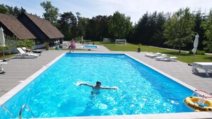 Una piscina rectangular con agua cristalina y azul, rodeada por un solárium con azulejos. Hay sillas de descanso y un aro de baloncesto en el solárium.