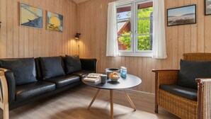 Una habitación acogedora con un sofá de cuero negro, un sillón de mimbre, una mesa redonda con un libro y una tetera, y paredes de madera con dos cuadros enmarcados.