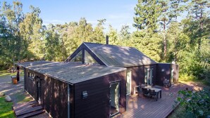En moderne hytte med terrasse, store vinduer og et skråt tag, omgivet af skov.