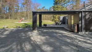 Et moderne hus med en overdækket terrasse, et legeområde og et stort træ i baggrunden.