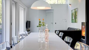 Et moderne spiseområde med et langt hvidt bord, gennemsigtige stole, en stor hængende lampe og en pejs.