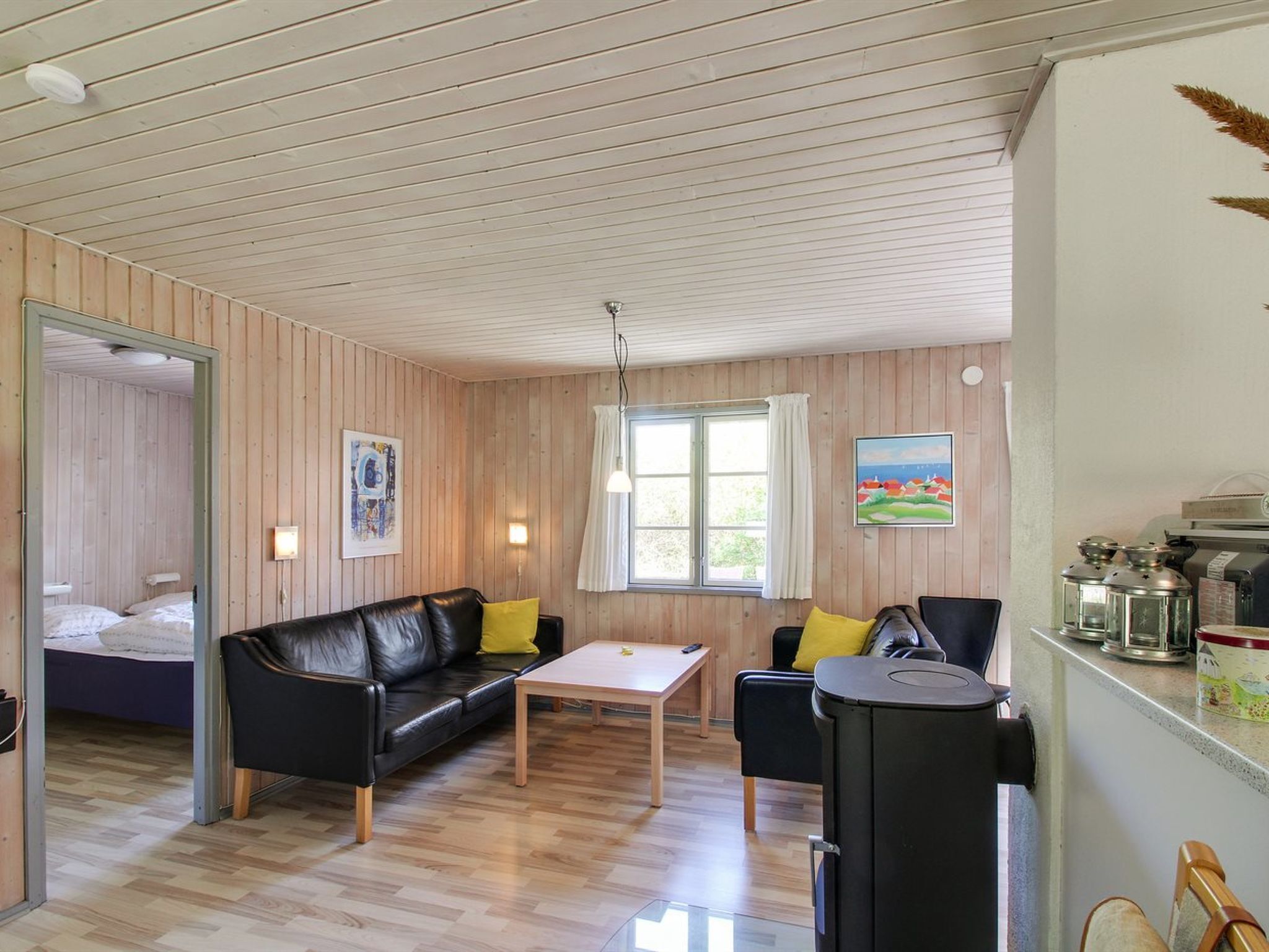 A cozy room with a wooden ceiling, a black leather sofa with yellow cushions, a wooden coffee table, and a kitchen area with a stove and jars.