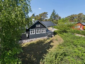 A black wooden cabin with a stone foundation, a small porch, and a red building in the background. - "Kerstin" - 100m from the sea by Interhome (Nexø)