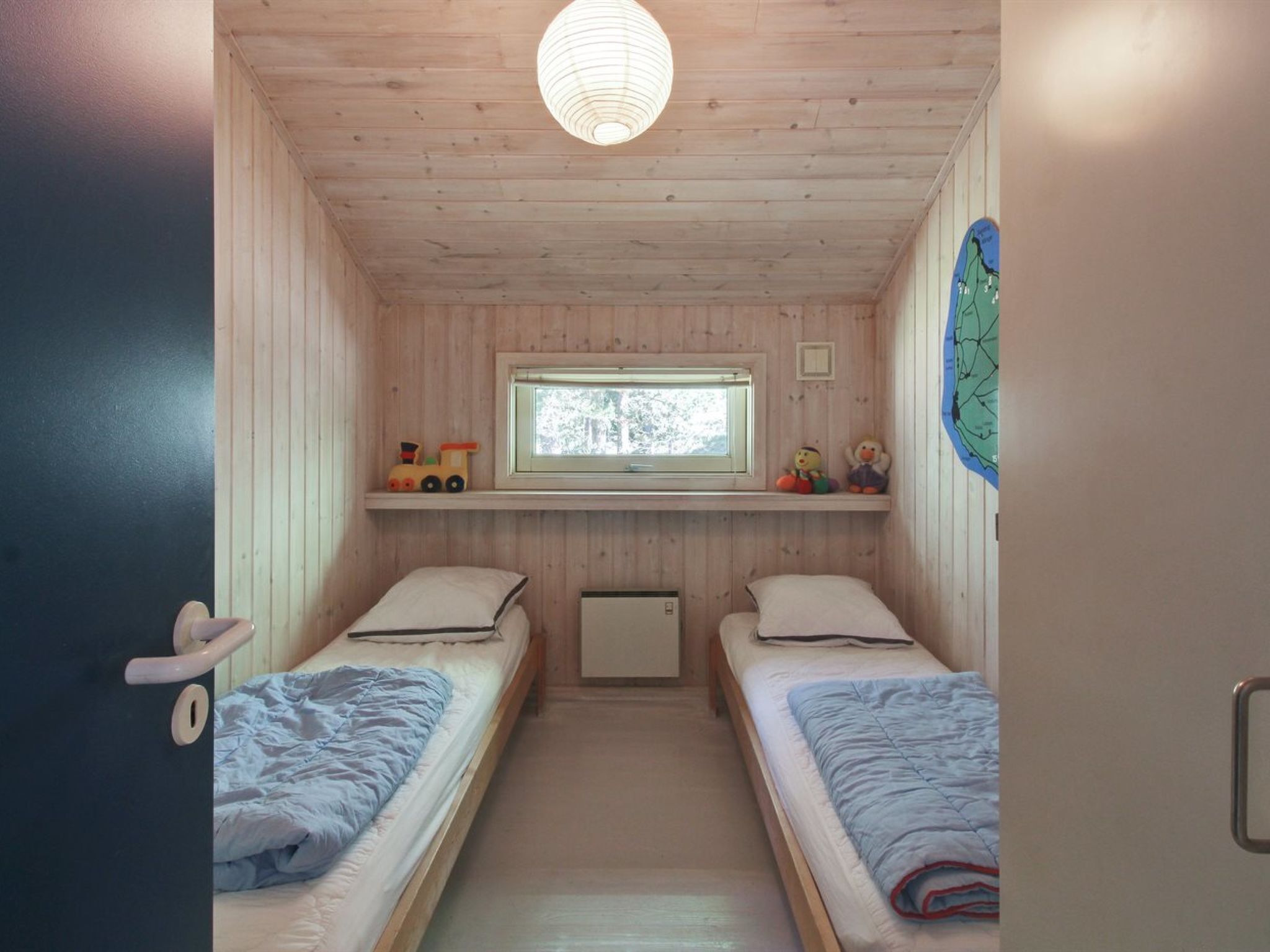 A compact bedroom with two single beds, a window, a shelf with toys, and a map on the wall.
