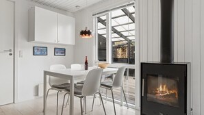 Ett modernt kök med en central köksö, vita skåp, en svart vedeldad spis och en matplats med ett bord och stolar med glasskiva.