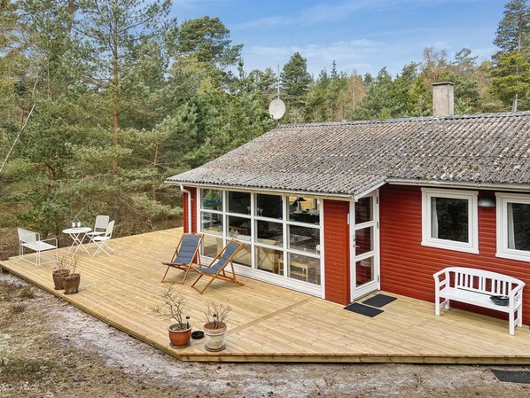Une terrasse en bois avec des meubles d’extérieur, un bâtiment rouge doté de grandes fenêtres et un paysage boisé en arrière-plan.
