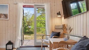 Una habitación acogedora con detalles de madera, un ventanal corredero que da a un jardín, una mesa con sillas, un televisor montado en la pared y un pequeño farol negro en el suelo.