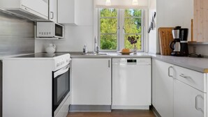 Una cocina moderna con armarios blancos, electrodomésticos de acero inoxidable, un ventanal con vistas a zonas verdes e iluminación colgante.
