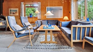 A cozy living room with wooden walls, a fireplace, comfortable seating, and large windows offering a view of the outdoors. - "Nada" - 200m from the sea by Interhome (Aakirkeby)