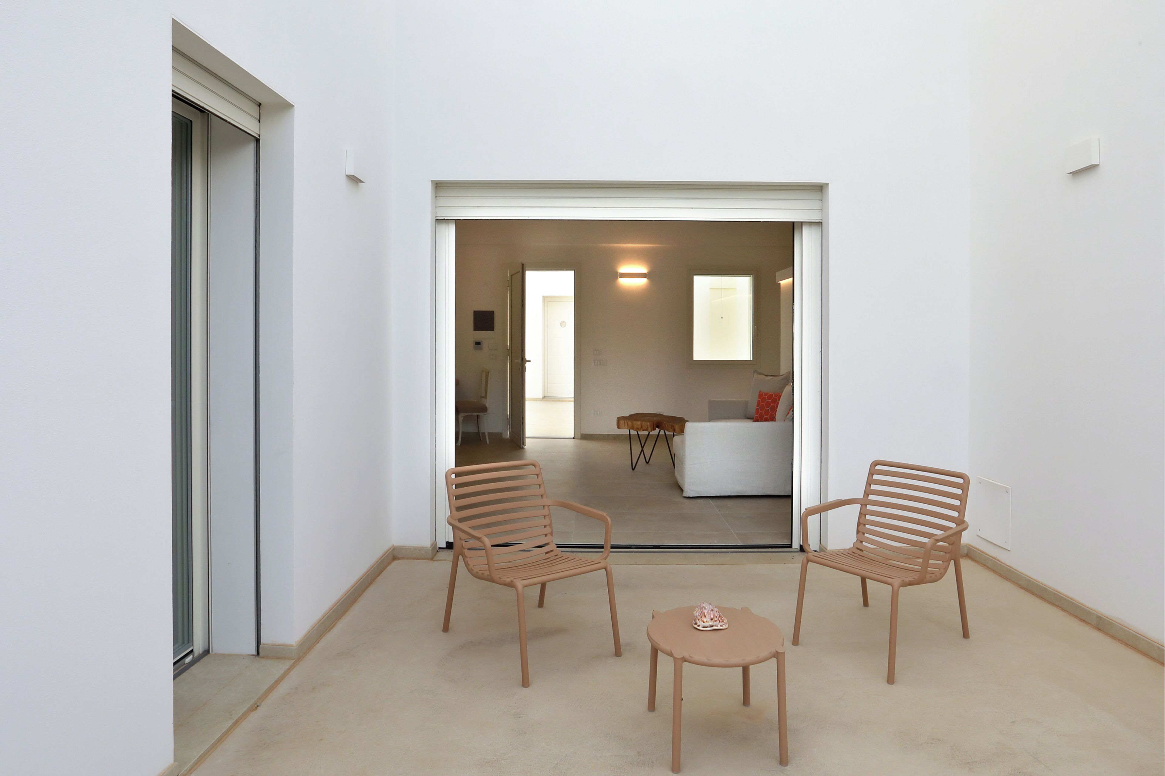 Un interior moderno con dos sillas de madera y una pequeña mesa, un sofá blanco y una puerta de vidrio que da a una sala de estar.