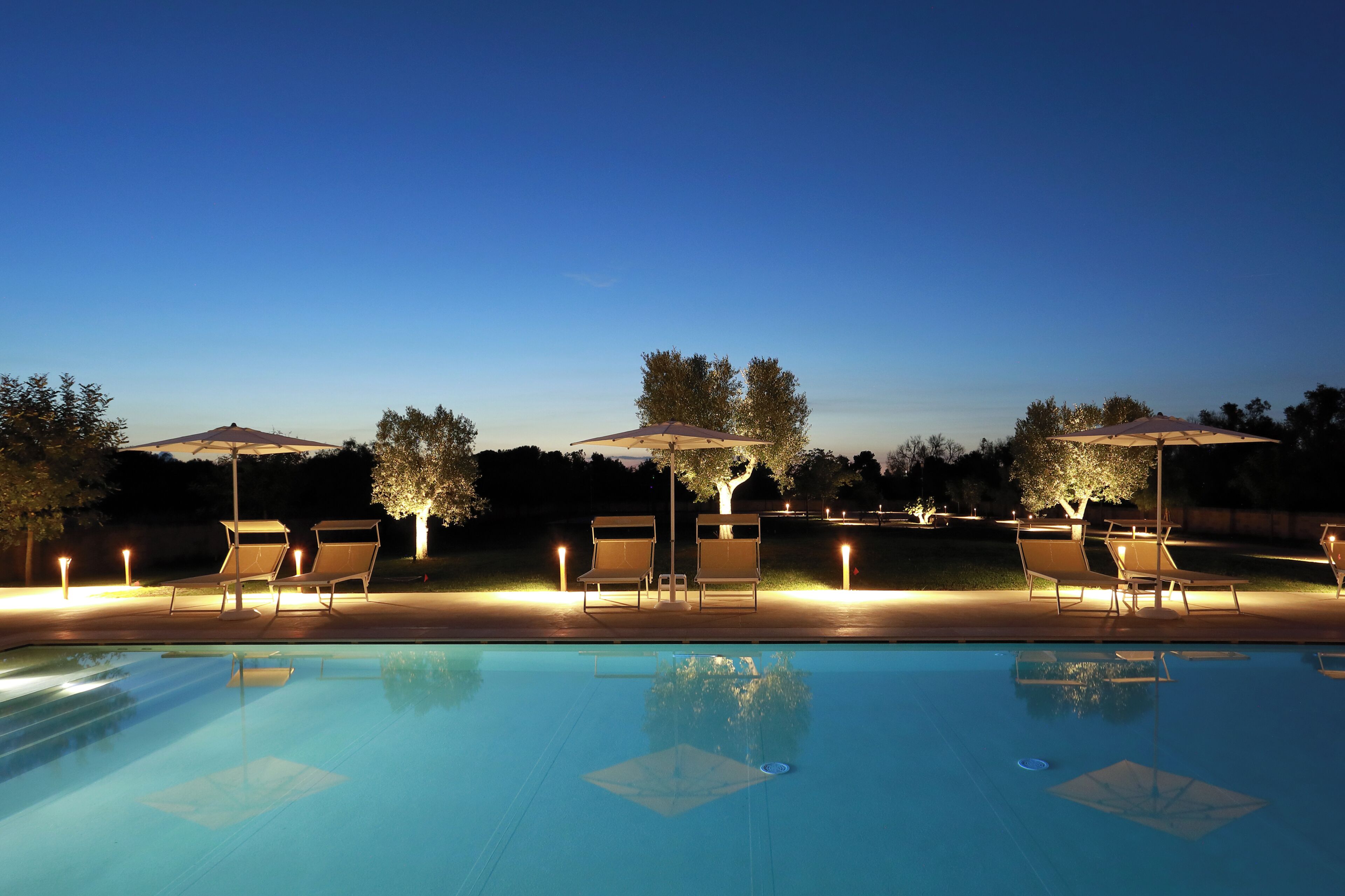 A swimming pool with lounge chairs and umbrellas, surrounded by trees and night lighting.