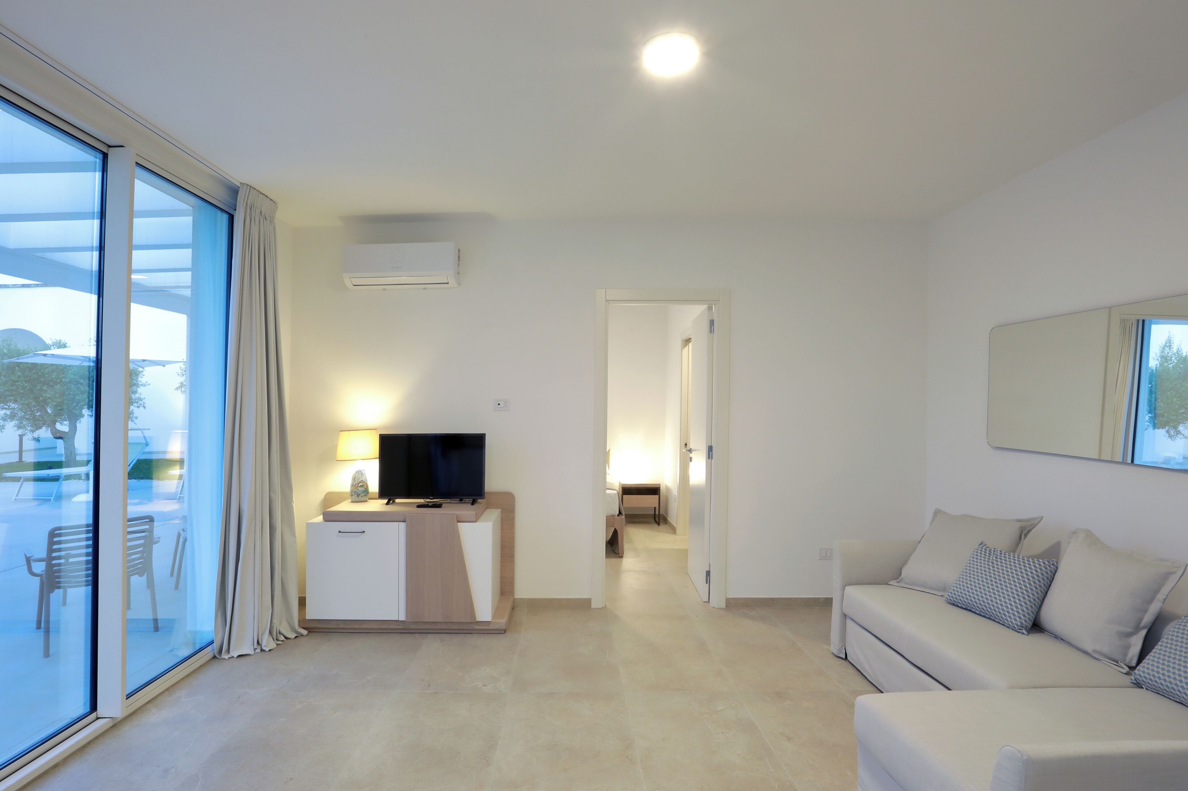 A modern living room with a sofa, a TV on a stand, and a glass door leading to an outdoor area.