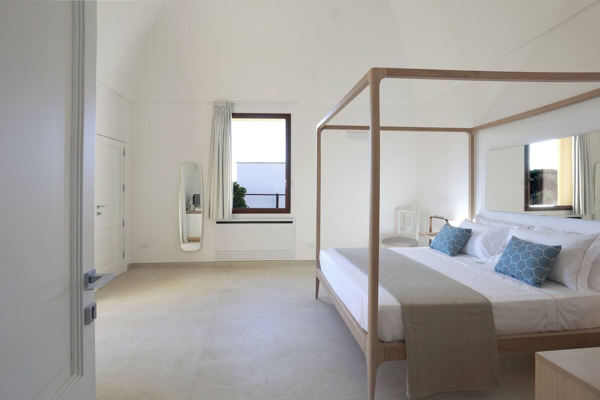 A bedroom with a four-poster bed, a window with curtains, a bedside table, and a mirror.