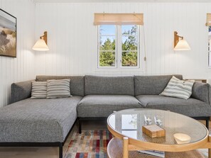 A modern living room with a grey sectional sofa, a glass coffee table, and a view of the outdoors through a window with a bamboo blind.