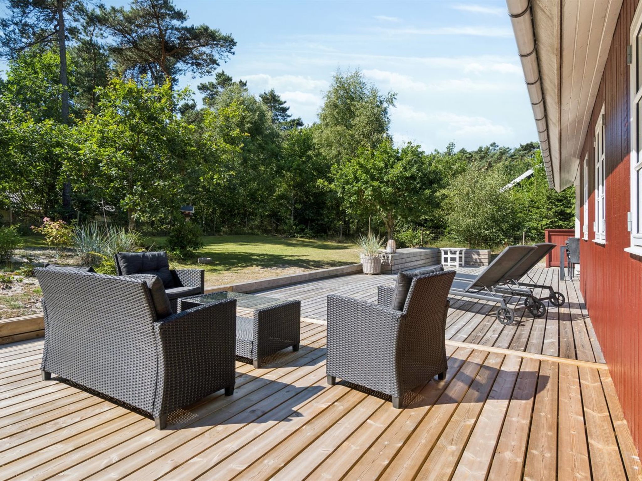 En træterrasse med udendørs møbler, en græsplæne og en rød bygning i baggrunden.
