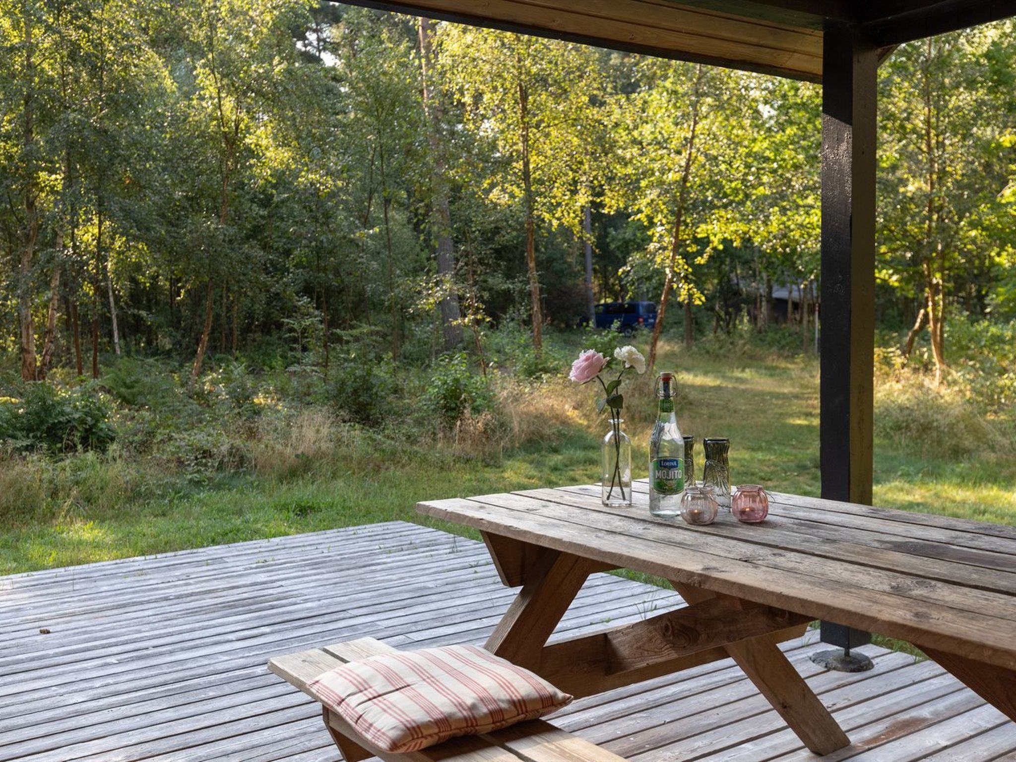 Ett picknickbord i trä med en kudde, en flaska sprit och en vas med blommor.